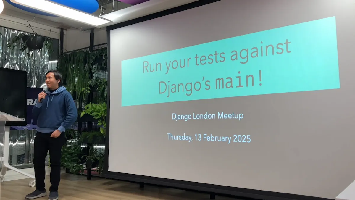 Sage presenting, holding a microphone. To his right his title slide states 'Run your tests against Django’s main!', 'Django London Meetup', 'Thursday, 13 February 2025'