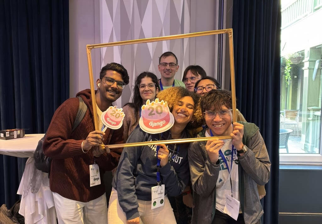 Seven people standing in a tight group behind a golden picture frame, smiling at the camera. Two hold birthday cake props that say 'Django' and '20'