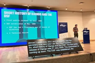Chaim Kirby on stage at DjangoCon Europe 2025 in Porto. He stands to the right of the stage next to the lectern, with a slide to his left 'Short history of Django and the DSF'. At the foreground there is a caption screen