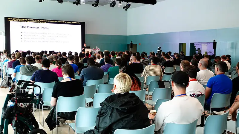 Conference talk about htmx, seen from the back of the room, with attendees sitting in rows in the foreground