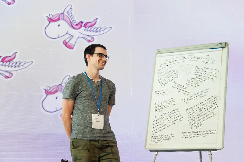 Mariusz on stage, smiling, next to a whiteboard that says 'Thank you Mariusz for your service'