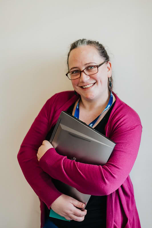 Rachell, smiling, wearing a black shirt and mauve top, holding her laptop