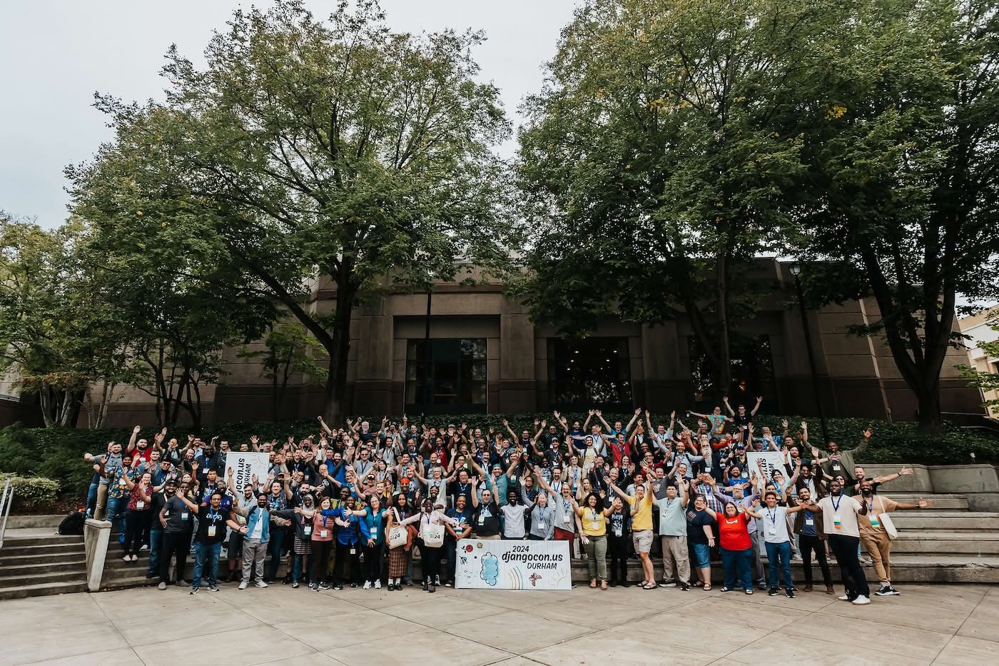 Group photo at DjangoCon US 2024, with all participants raising their arms while standing on stairs in front of the conference venue building