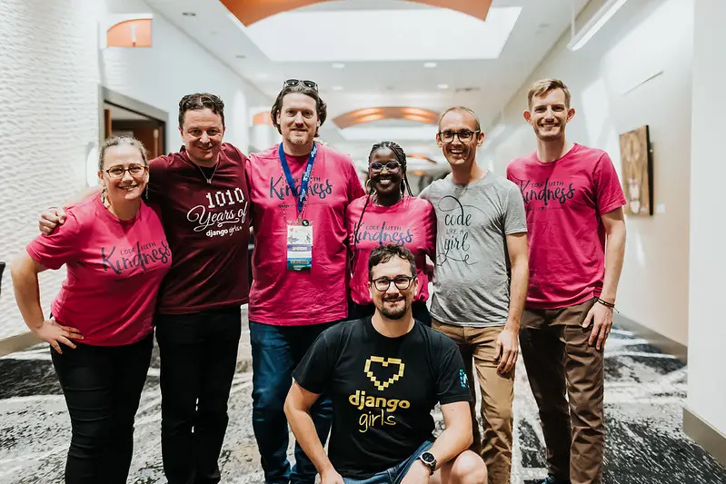 Seven Django community members wearing Django Girls+ shirts: Code with Kindness, Django Girls heart, 1010 years of Django Girls+, Code like a girl