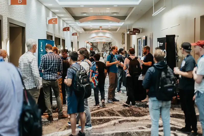 Hallway of DjangoCon US venue, with attendees talking to one-another in a few groups, and sponsor tables to the sides