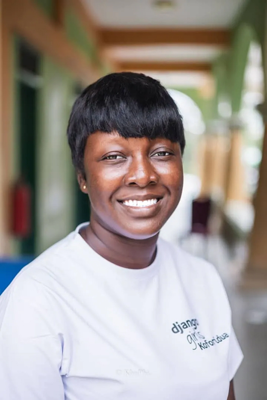 Theresa portrait, a pretty black woman with short hair. She is looking at the camera with a big smile. She wears a white t-shirt with written in green "Django Girls Koforidua". The background is blurry but really colorful with shades of green and orange.