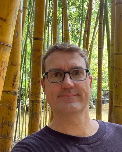 Rob Hudson's profile, with bamboos behind him. He wear glasses and slightly smiling in front the camera.