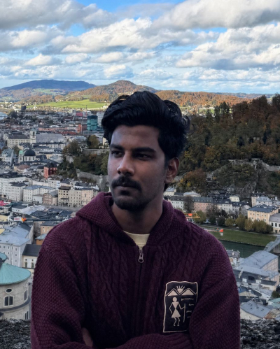 Bhuvnesh, with a dark red pull over, in front of the camera, looking on the left. We can see a landscape with multiple buildings behind him and the cloudy sky.