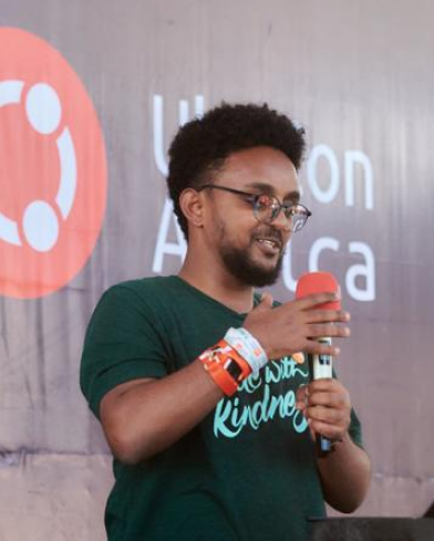 Salim, wears black glasses and a green t-shirt written "code with kindness". He is speaking at DjangoCon Africa, with a mic in one hand and smiling while looking at his computer.