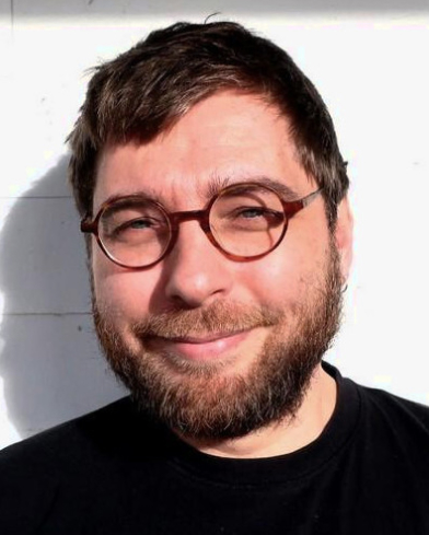 Benjamin's profile, smiling with his glasses to the camera. He wears a black top and the background is white.